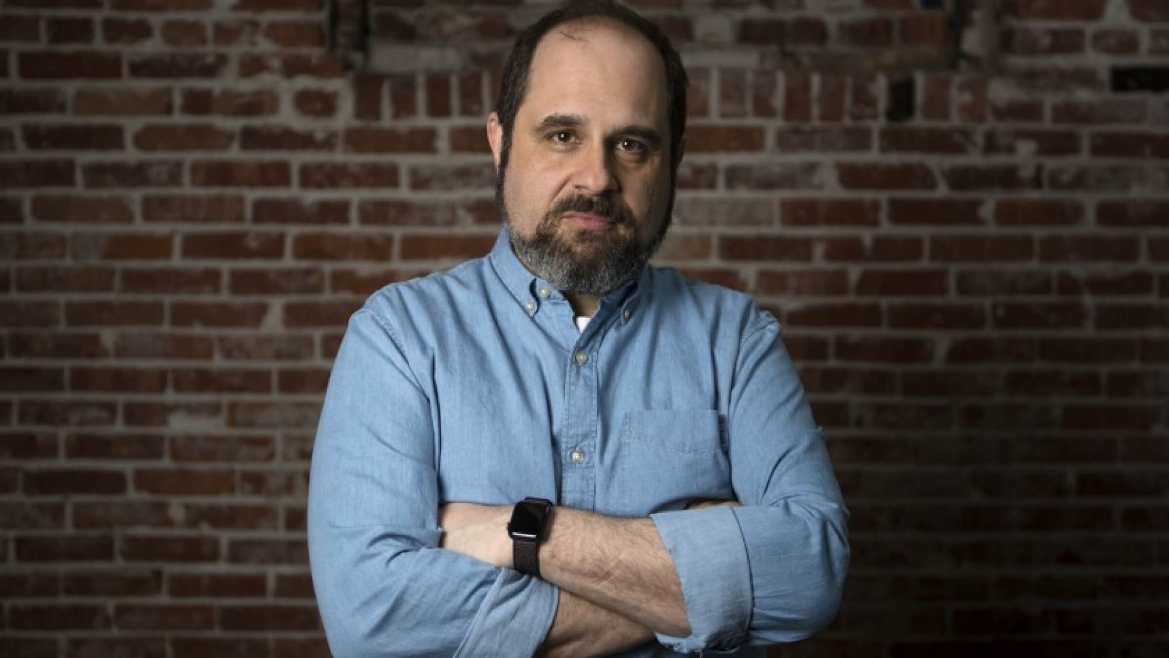 Craig Mazin menggunakan kemeja warna biru dan jam tangan hitam, ia melipatkan tangannya ke dada